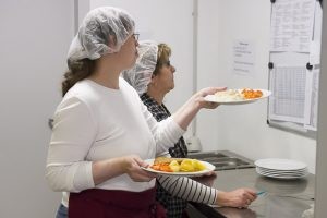 Mitarbeiterinnen des DFG Hagen schauen auf eine Liste. Eine Mitarbeiterin hält bereits das fertige Essen in den Händen.