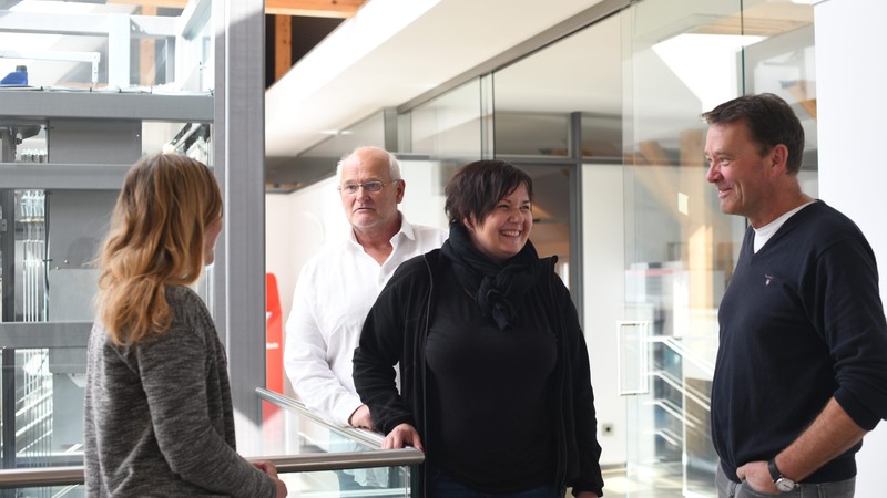 Jochen Twelker (hinten links), Heidi Emmerich und Martin Kapovits (vorne rechts) stehen gemeinsam in einem großen Flur und unterhalten sich.