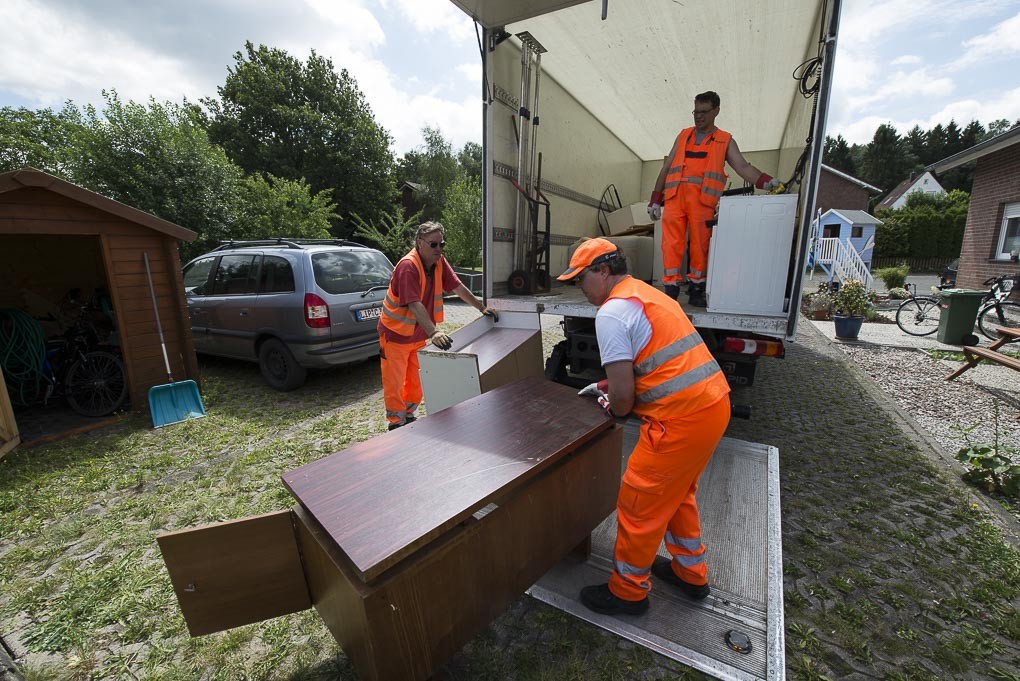 Drei Mitarbeiter der AGA in orange-farbenen Westen laden einen Schrank vom Sperrmüll in ihren Lieferwagen.
