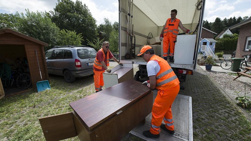 Drei Mitarbeiter der AGA in orange-farbenen Westen laden einen Schrank vom Sperrmüll in ihren Lieferwagen.